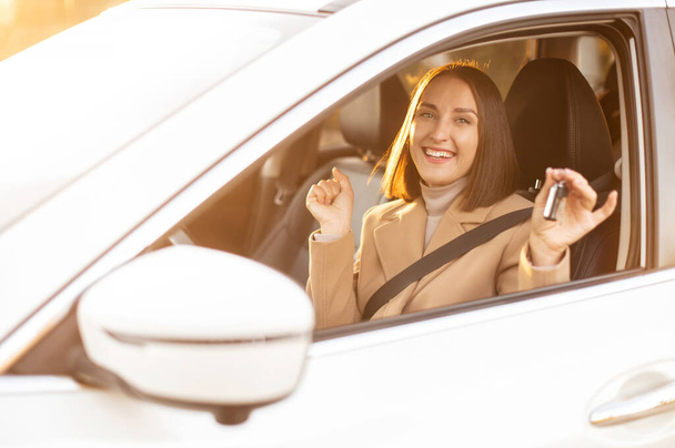 Cheerful woman showing key from new car, sitting inside looks at the camera and smiling, successful female buying a car - Fotó, kép