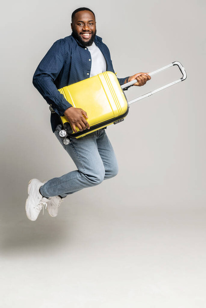 Joyful African-American guy jumping with a yellow suitcase, plays on a bag like a guitarist, fooling around, a black man with luggage looking forward a trip, isolated on gray, full length, vertical - Foto, Imagen