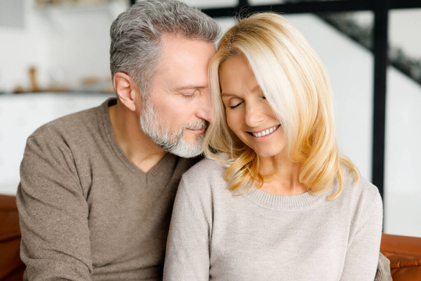 Cheerful and happy middle-aged spouses enjoying time together. Handsome grey-haired man hugging gently beautiful good-looking woman, their eyes are closed. Love and bond concept. Close-up portrait - 写真・画像