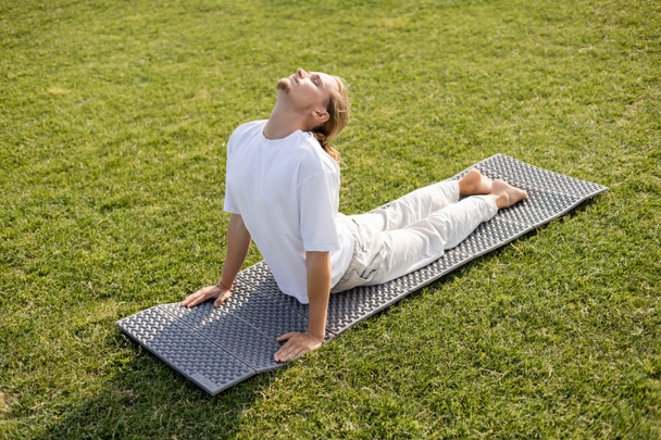 Beyaz tişörtlü ve keten pantolonlu çıplak ayaklı bir adam yeşil çimlerde kobra pozisyonunda yoga yapıyor.  - Fotoğraf, Görsel