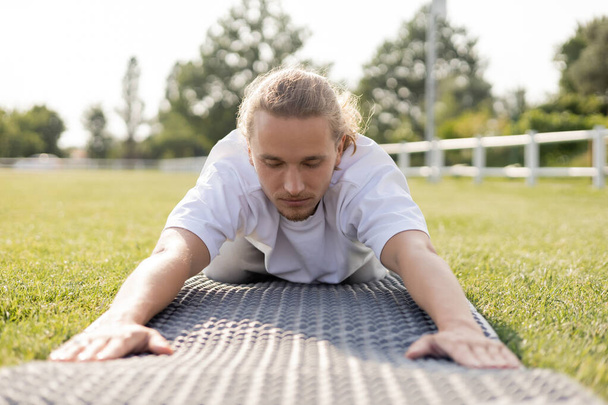 Oberflächenebene eines jungen Mannes, der mit geschlossenen Augen auf einer Yogamatte auf grünem Rasen meditiert - Foto, Bild