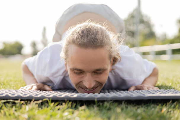 nivel de superficie del hombre despreocupado con los ojos cerrados sonriendo mientras practica yoga sobre hierba verde al aire libre - Foto, Imagen