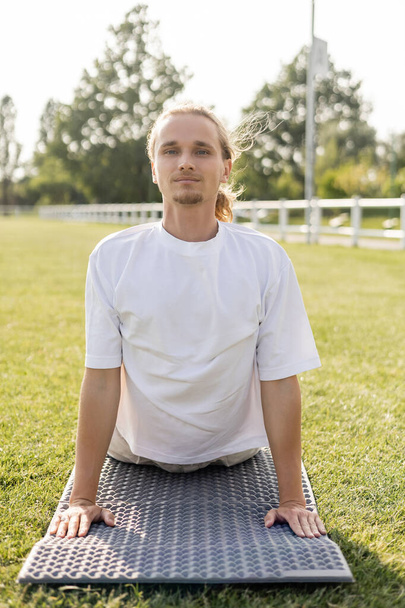 jong positief man in wit t-shirt kijken naar camera tijdens het oefenen cobra pose op yoga mat buiten - Foto, afbeelding