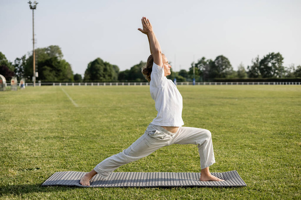 side view of sportive barefoot man meditating in warrior pose with raised hands on stadium outdoors - Photo, image