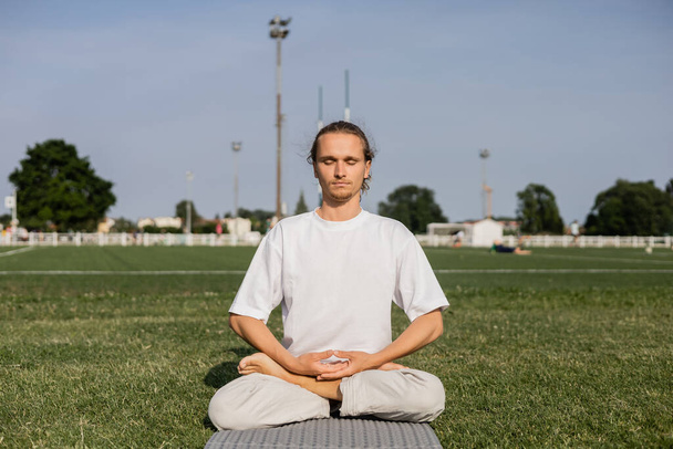 молодой человек в белой футболке медитирует в позе лотоса с закрытыми глазами на зеленом поле открытого стадиона - Фото, изображение