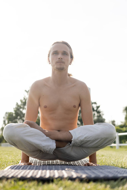 joven hombre sin camisa en pantalones de lino practicando yoga a escala posan sobre esterilla de yoga al aire libre - Foto, Imagen