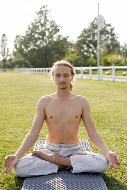 hombre sin camisa en pantalones de lino meditando en pose de loto mientras está sentado en la esterilla de yoga con los ojos cerrados al aire libre - Foto, Imagen