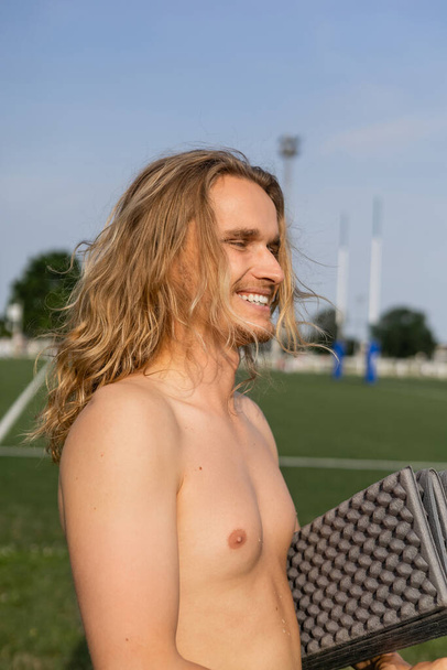 nadšený muž bez košile s dlouhými vlasy drží jógu rohožku a usmívá se venku - Fotografie, Obrázek