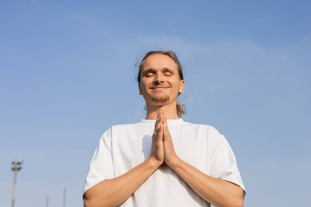 potěšený muž v bílém bavlněném tričku meditující se zavřenýma očima a anjali mudra gestem proti jasné obloze - Fotografie, Obrázek