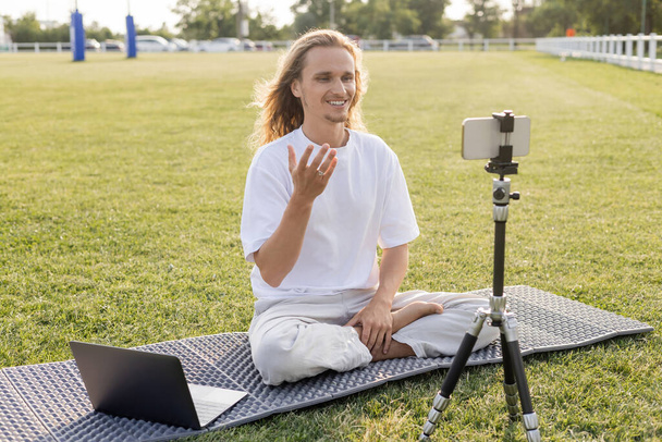 entrenador de yoga feliz sentado en pose fácil cerca del ordenador portátil y el teléfono móvil en el trípode en el césped verde del estadio al aire libre - Foto, Imagen