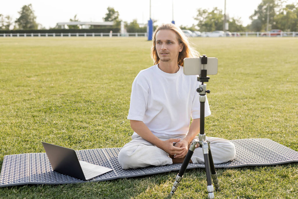 long haired yoga teacher looking away while sitting near laptop and tripod with mobile phone on field of outdoor stadium - Foto, afbeelding