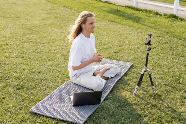 mosolygós jóga tanár ül könnyű pózban közel állvány okostelefon és laptop zöld gyep - Fotó, kép