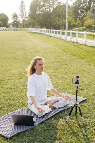 yoga teacher showing easy pose and gyan mudra gestures while sitting on yoga mat near smartphone on tripod outdoors - Photo, Image