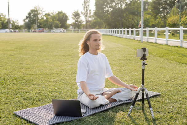 yoga vlogger meditando en pose fácil cerca del teléfono móvil en trípode en césped verde del estadio al aire libre - Foto, Imagen