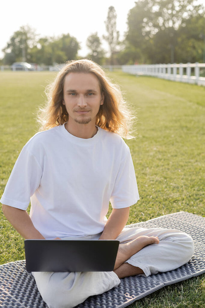 positieve langharige yoga man zitten in gemakkelijke pose in de buurt laptop en kijken naar de camera op groen gazon - Foto, afbeelding