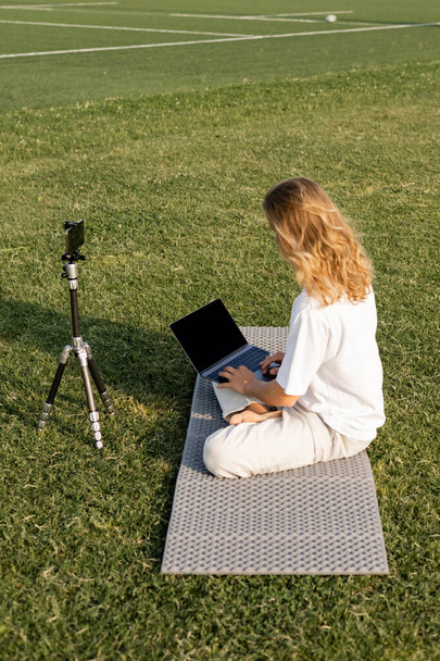 long haired yoga teacher using laptop while sitting near tripod with cellphone on grassy stadium - Photo, Image