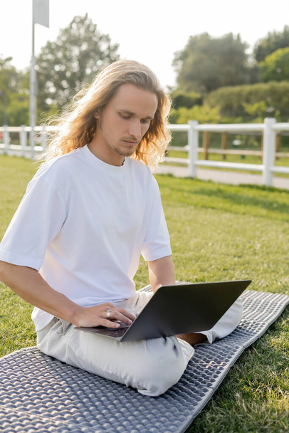 若いヨガの男は芝生のスタジアムで簡単なポーズで座ってオンラインレッスン中にラップトップを使用して - 写真・画像