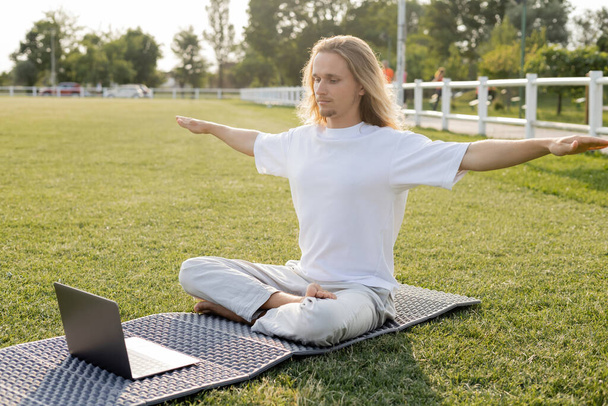 молодий чоловік сидить у легкій позі з розтягнутими руками під час онлайн-урок йоги на ноутбуці на відкритому повітрі
 - Фото, зображення