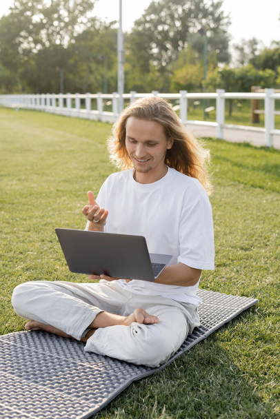 улыбающийся мужчина разговаривает во время урока йоги на ноутбуке, сидя в легкой позе на открытом стадионе - Фото, изображение