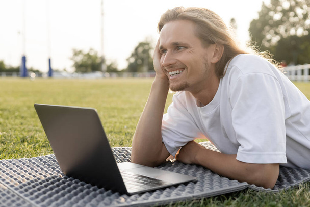 young cheerful yoga man looking away while lying near laptop on grassy stadium outdoors - Photo, Image