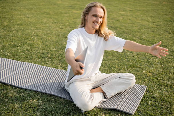 homme heureux avec ordinateur portable montrant geste de salutation tout en étant assis sur tapis de yoga sur la pelouse verte à l'extérieur - Photo, image