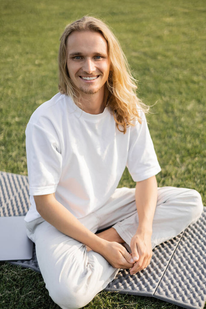 carefree man sitting on yoga mat near laptop and smiling at camera outdoors - Foto, Bild