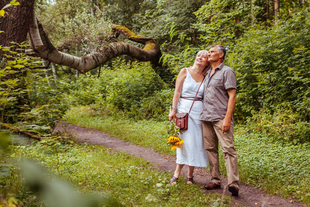 Κομψό ζευγάρι ηλικιωμένων που περπατούν έξω στο καλοκαιρινό δάσος. Ηλικιωμένοι κρατιούνται χέρι χέρι. Η γυναίκα κουβαλάει μπουκέτο λουλούδια. Άνδρας και γυναίκα απολαμβάνουν το τοπίο - Φωτογραφία, εικόνα