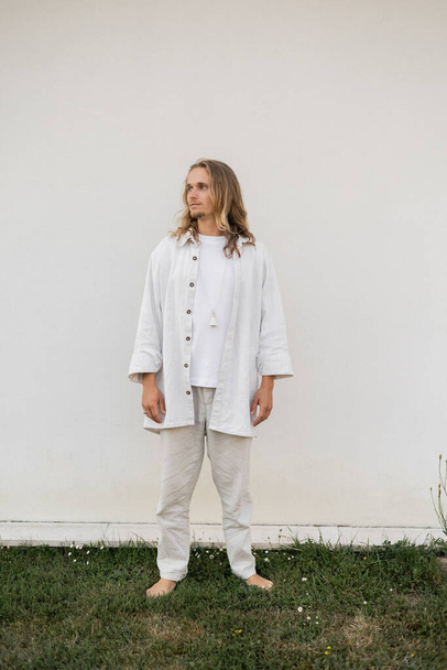 full length of young barefoot man in white linen clothes standing near wall outdoors and looking away - Fotó, kép