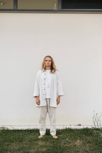 longitud completa de hombre descalzo y de pelo justo en ropa de lino de pie cerca de la pared blanca al aire libre - Foto, imagen