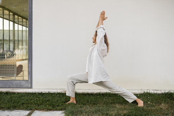 side view of barefoot man in white cotton clothes meditating in warrior pose with raised praying hands near modern cottage - Fotografie, Obrázek