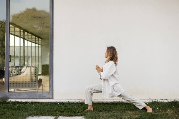 vista lateral del hombre de pelo largo en ropa de lino meditando en pose guerrera con las manos de oración cerca de la pared blanca de la casa de campo moderna - Foto, Imagen
