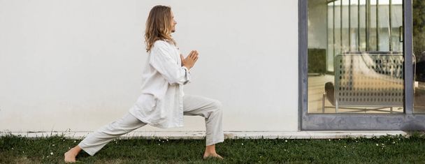 vista laterale di uomo elegante in abiti bianchi praticare yoga in posa guerriero con gesto mudra anjali vicino casa moderna, banner - Foto, immagini