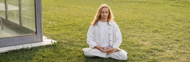 Beyaz elbiseli genç adam Lotus 'ta meditasyon yapıyor. Binanın yanındaki yeşil çimlerde poz veriyor. - Fotoğraf, Görsel