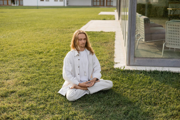 stylish long haired man meditating in lotus pose while sitting on grassy lawn near building - Photo, Image