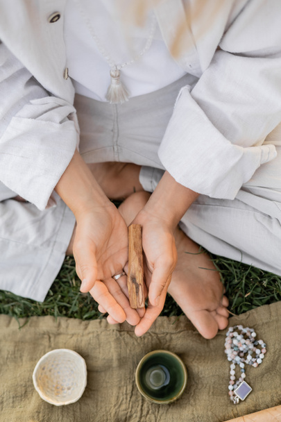 vue du dessus de l'homme cultivé en vêtements blancs tenant palo santo bâton près du tapis de lin avec des tasses en céramique et des perles de mala  - Photo, image
