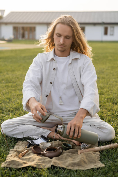 long haired man in linen clothes pouring hot water from thermos into ceramic teapot  - Foto, afbeelding