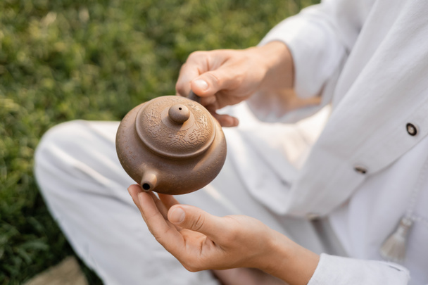 cropped view of man in white linen clothes holding clay teapot while sitting outdoors - Photo, Image