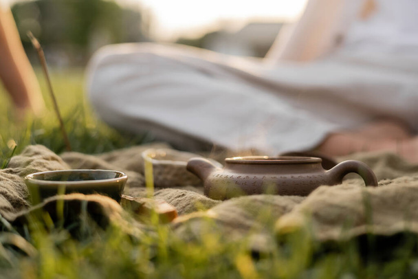 linen rug with ceramic teapot and cups on green grass near man in blurred background  - Photo, Image