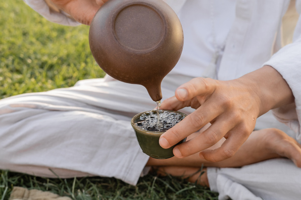 partial view of yoga man in linen clothes sitting on green lawn and pouring puer tea from oriental teapot in clay bowl  - Fotografie, Obrázek