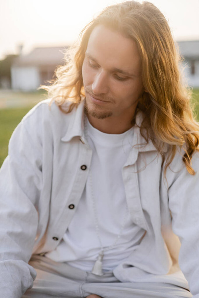 young and dreamy man with long fair hair meditating while sitting outdoors - Photo, Image
