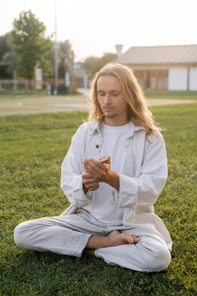 stylish long haired man in white clothes meditating in easy pose and holding palo santo stick on green lawn - Foto, Imagem