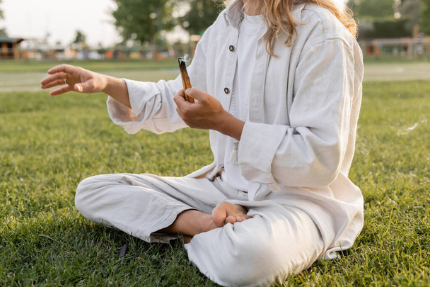 visão parcial do homem em roupas brancas segurando palo ardente santo vara e meditando em pose fácil no gramado verde - Foto, Imagem