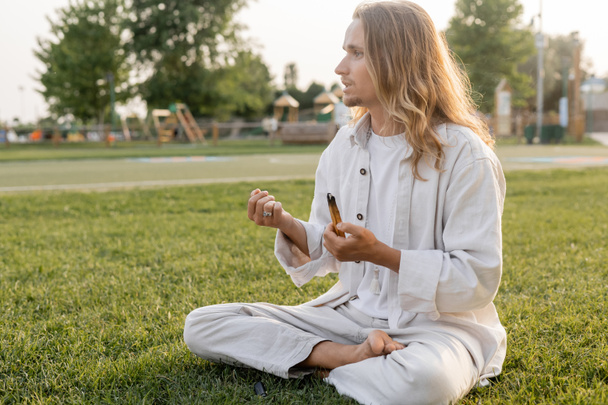 Barfuß-Mann in weißen Leinenkleidern meditiert in lockerer Pose mit schwelendem Palo Santo Stick und schaut draußen weg - Foto, Bild