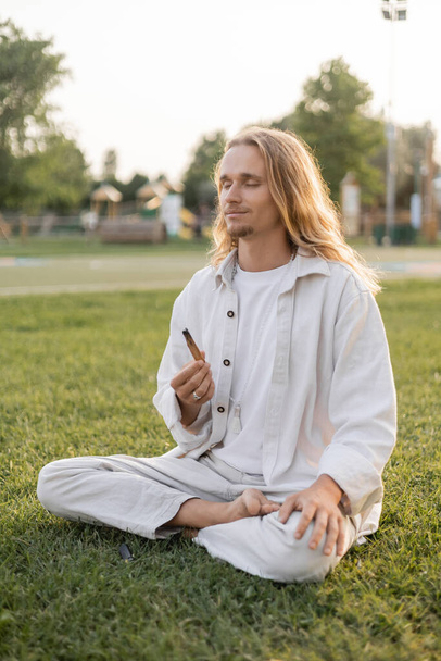 pleased man in white clothes holding aromatic palo santo stick during meditation in sukhasana pose on green grass  - Fotó, kép