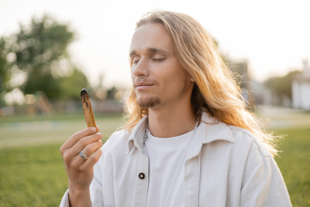 pleased long haired man with closed eyes holding smoldering palo santo stick while meditating outdoors - 写真・画像