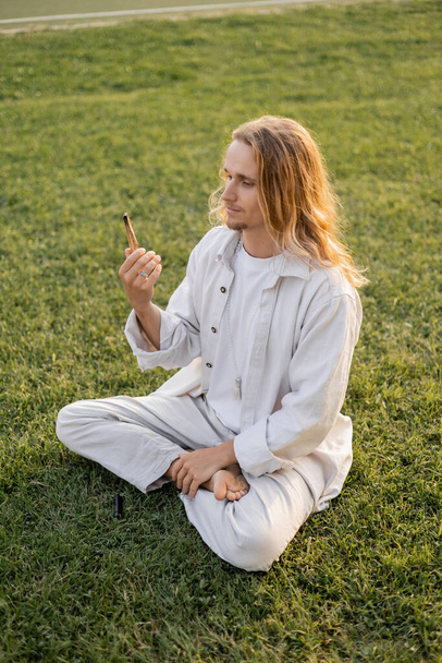 young yoga man in white clothes looking at fragrant palo santo stick while sitting in sukhasana pose on green grass - Fotoğraf, Görsel