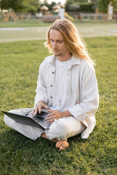 młody długowłosy mężczyzna w białych bawełnianych ubraniach siedzi w łatwej pozie i za pomocą laptopa na zielonym trawniku - Zdjęcie, obraz