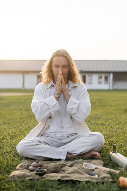young man in white clothes meditating in easy pose near linen rug with clay teapot and bowls on green lawn - Photo, Image