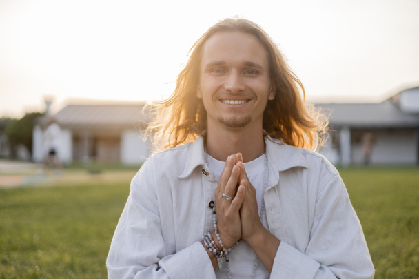 carefree long haired man with mala beads showing anjali mudra gesture and smiling at camera outdoors - Photo, Image