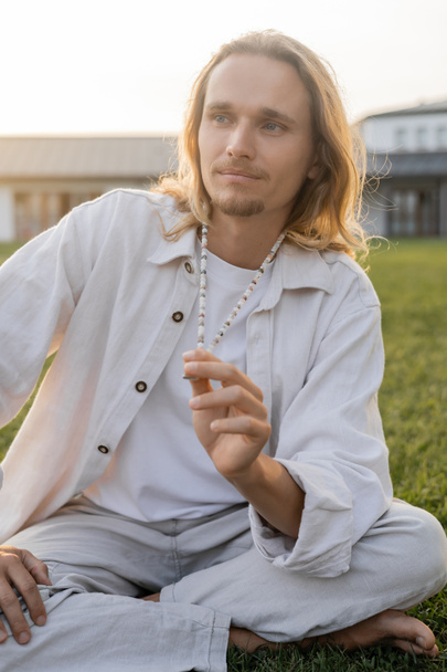 long haired man in linen clothes touching mala beads and looking away during meditation outdoors - Foto, Bild